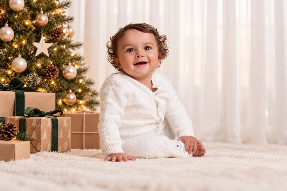 A baby on a cream rug beside a Christmas tree